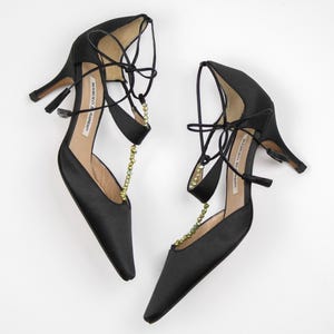 Puede incluir: Un par de zapatos de tac&oacute;n alto de sat&eacute;n negro con punta. Los zapatos tienen delicadas correas en el tobillo y est&aacute;n adornados con una cadena de peque&ntilde;as cuentas doradas. Los tacones miden aproximadamente 7,6 cm de alto.