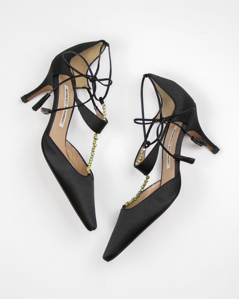 Puede incluir: Un par de zapatos de tac&oacute;n alto de sat&eacute;n negro con punta. Los zapatos tienen delicadas correas en el tobillo y est&aacute;n adornados con una cadena de peque&ntilde;as cuentas doradas. Los tacones miden aproximadamente 7,6 cm de alto.