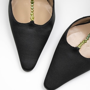 Puede incluir: Un par de zapatos de sat&eacute;n negro con punta. Los zapatos tienen una delicada correa adornada con peque&ntilde;as cuentas verdes. El interior de los zapatos es de color beige claro, sobre un fondo blanco.