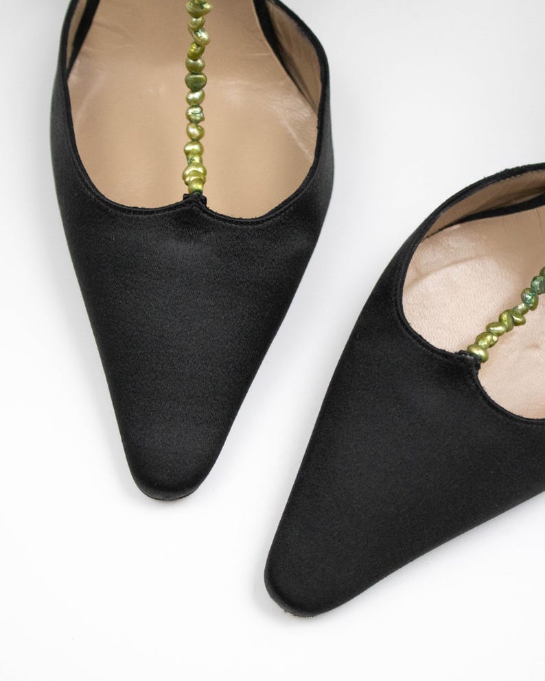 Puede incluir: Un par de zapatos de sat&eacute;n negro con punta. Los zapatos tienen una delicada correa adornada con peque&ntilde;as cuentas verdes. El interior de los zapatos es de color beige claro, sobre un fondo blanco.