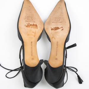 Puede incluir: Un par de tacones Manolo Blahnik de sat&eacute;n negro, talla 36, con puntera y lazos en el tobillo. Las suelas son de color marr&oacute;n claro con el nombre de la marca y la talla impresos. Los tacones tienen una peque&ntilde;a borla al final del lazo.