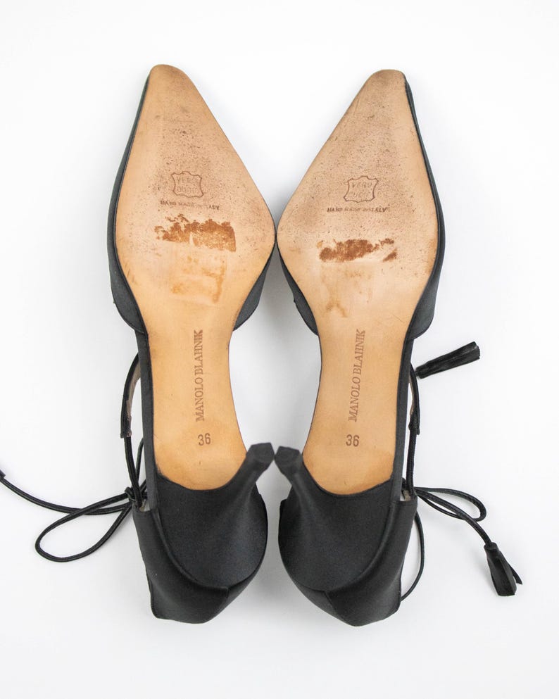 Puede incluir: Un par de tacones Manolo Blahnik de sat&eacute;n negro, talla 36, con puntera y lazos en el tobillo. Las suelas son de color marr&oacute;n claro con el nombre de la marca y la talla impresos. Los tacones tienen una peque&ntilde;a borla al final del lazo.