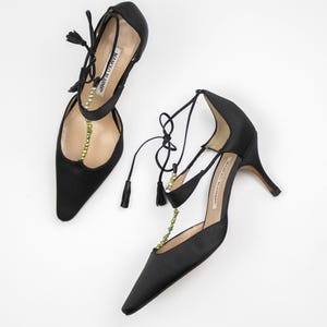 Puede incluir: Un par de zapatos de tac&oacute;n alto de sat&eacute;n negro con punta. Los zapatos presentan delicadas cintas en el tobillo con borlas y una cadena de peque&ntilde;as cuentas doradas. El nombre de la marca es visible en la plantilla.