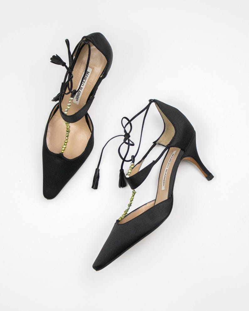 Puede incluir: Un par de zapatos de tac&oacute;n alto de sat&eacute;n negro con punta. Los zapatos presentan delicadas cintas en el tobillo con borlas y una cadena de peque&ntilde;as cuentas doradas. El nombre de la marca es visible en la plantilla.