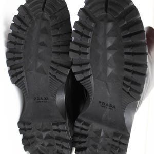 May include: Black Prada boots with a rugged, textured sole. The soles are thick with deep treads for traction. The brand name "PRADA" is embossed on each sole. The boots are likely made of leather or a similar material.
