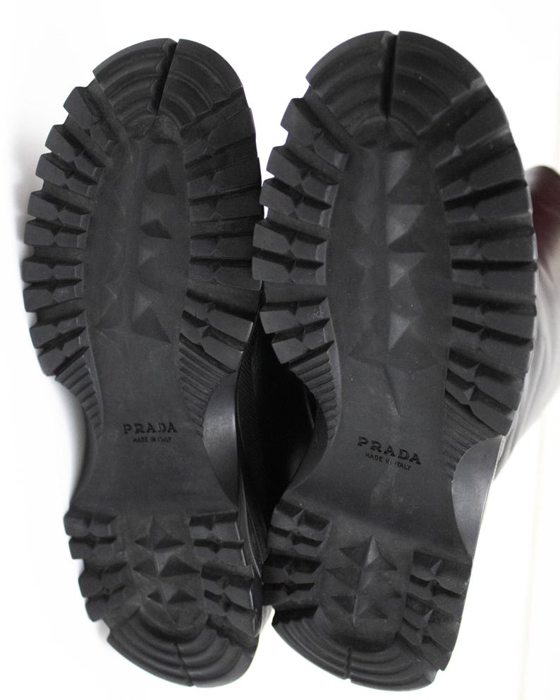 May include: Black Prada boots with a rugged, textured sole. The soles are thick with deep treads for traction. The brand name "PRADA" is embossed on each sole. The boots are likely made of leather or a similar material.