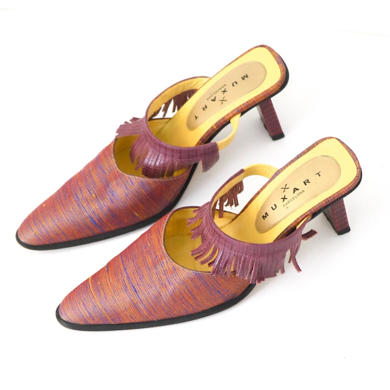 K&ouml;nnte beinhalten: Ein Paar violette und orangefarbene Mules mit Fransen. Die Schuhe haben eine spitze Zehenpartie und eine gelbe Innensohle mit dem aufgedruckten Text "MU*ART BARCELONA".