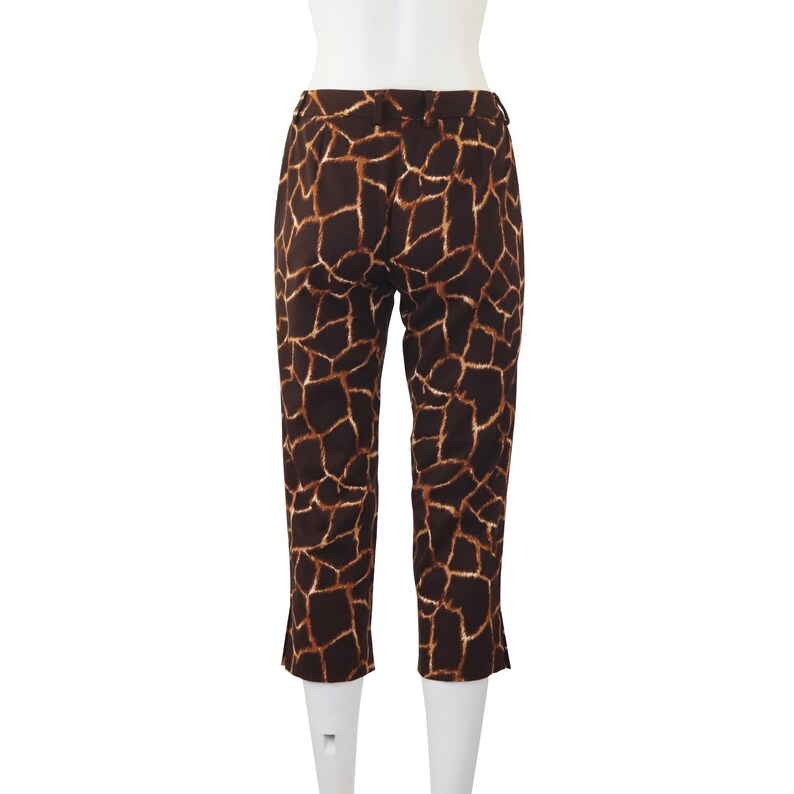 May include: Cropped trousers featuring a giraffe print in brown and tan. The back view of the trousers is displayed on a mannequin. The fabric has a repeating pattern of irregular shapes in varying shades of brown and tan.