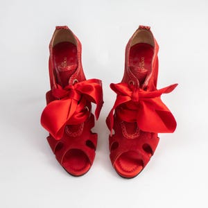 Puede incluir: Un par de tacones rojos de ante con la punta abierta y cordones de cinta de raso. Los tacones tienen un diseño de punta abierta y están sobre un fondo blanco. Los tacones son de color rojo intenso.
