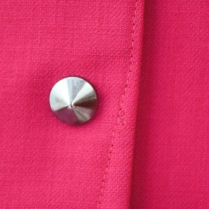 May include: A close-up of a silver button on a bright pink fabric. The button is round and has a pointed top.