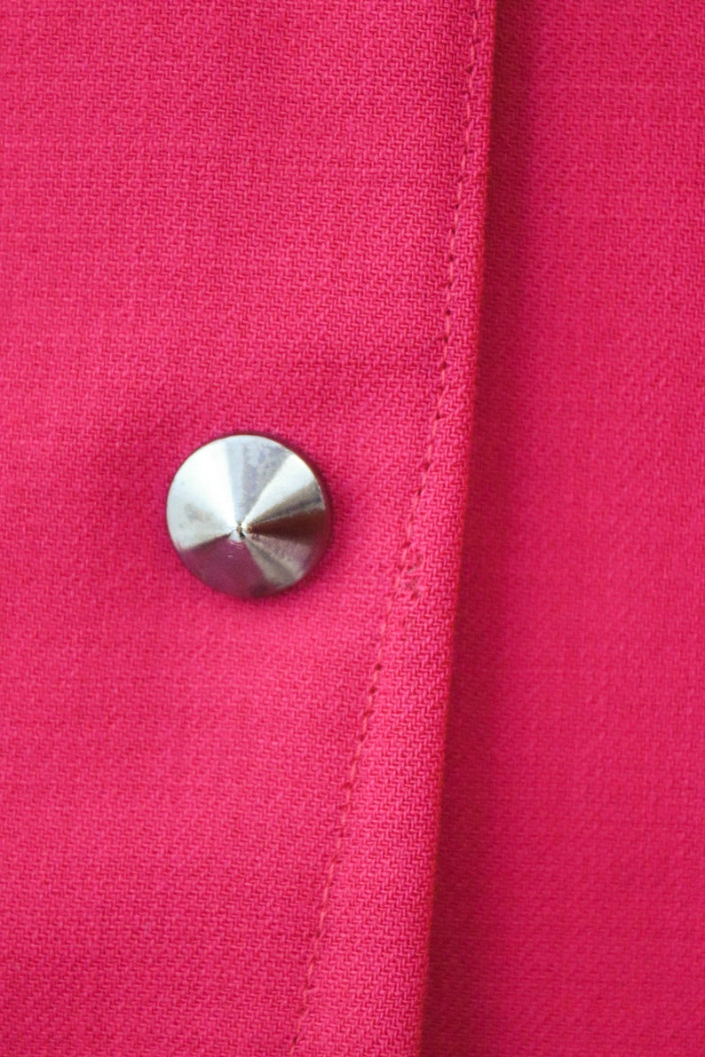 May include: A close-up of a silver button on a bright pink fabric. The button is round and has a pointed top.