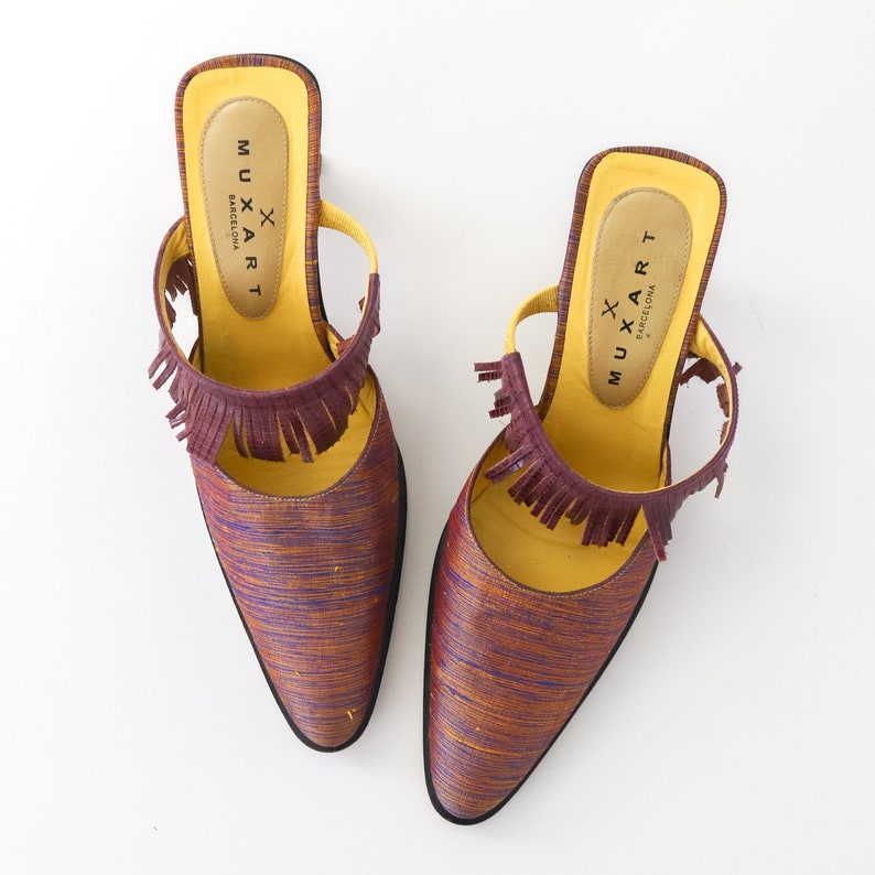 K&ouml;nnte beinhalten: Ein Paar violette und gelbe spitze Mules mit Fransen. Die Schuhe haben eine gelbe Lederinnensohle mit dem aufgedruckten Text "MUXART BARCELONA".