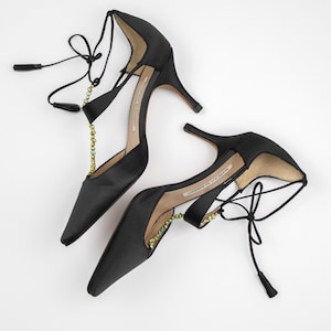 Puede incluir: Un par de zapatos de tac&oacute;n alto de sat&eacute;n negro con punta. Los zapatos presentan delicadas cintas en el tobillo con peque&ntilde;as borlas y una cadena decorativa con cuentas verdes. Los tacones son delgados y elegantes.