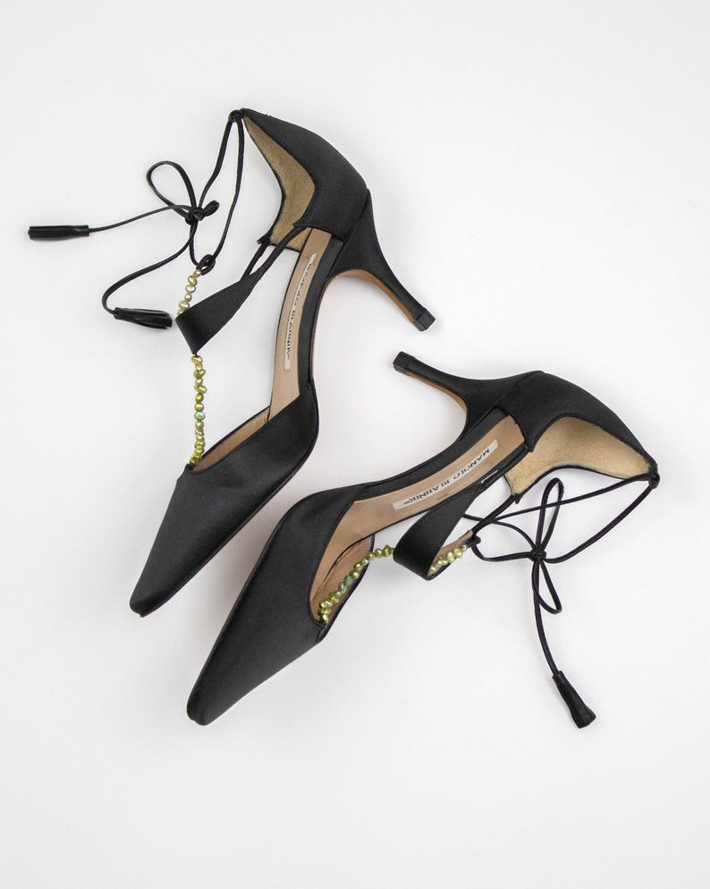 Puede incluir: Un par de zapatos de tac&oacute;n alto de sat&eacute;n negro con punta. Los zapatos presentan delicadas cintas en el tobillo con peque&ntilde;as borlas y una cadena decorativa con cuentas verdes. Los tacones son delgados y elegantes.