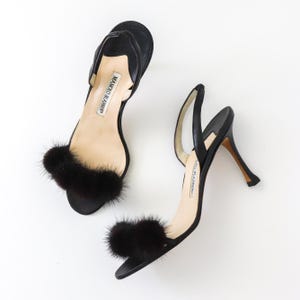 May include: A pair of black heeled sandals with black fur pom-poms on the straps. The sandals are made by Manolo Blahnik.
