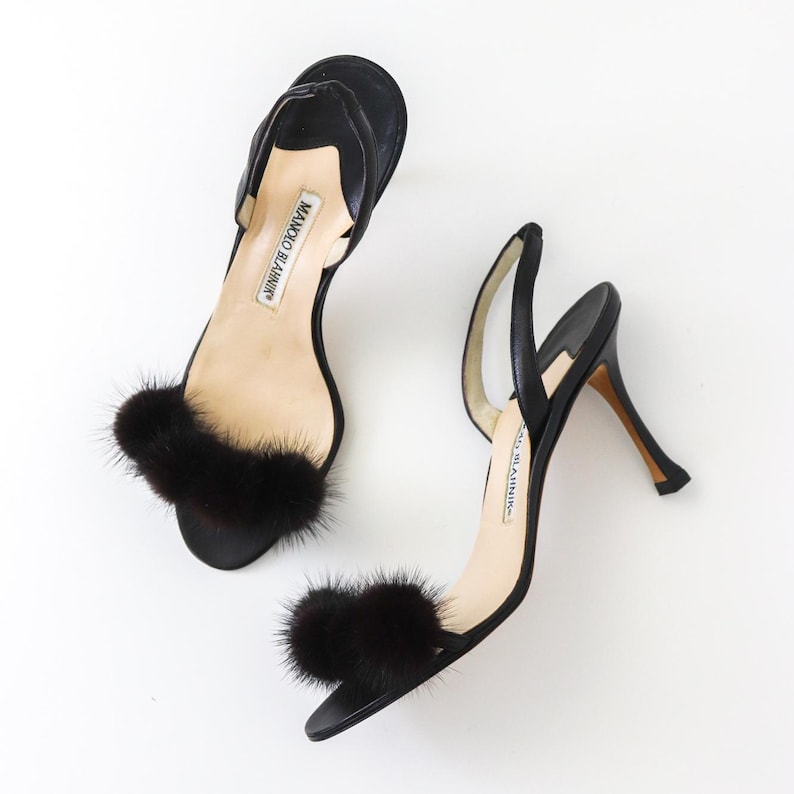 May include: A pair of black heeled sandals with black fur pom-poms on the straps. The sandals are made by Manolo Blahnik.