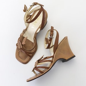 May include: A pair of brown leather wedge sandals with butterfly accents. The sandals have a square toe, ankle straps, and a wooden wedge heel. The brand name "Prada" is visible on the insole.