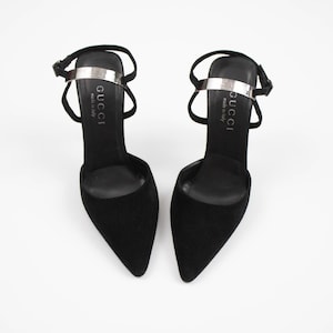 May include: A pair of black suede high-heeled shoes with pointed toes. Each shoe has a silver strap across the top with the brand name "GUCCI" printed on it. The shoes have thin ankle straps and are set against a white background.