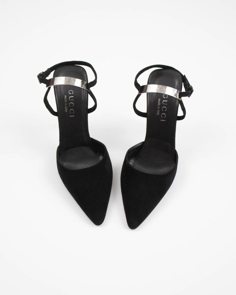 May include: A pair of black suede high-heeled shoes with pointed toes. Each shoe has a silver strap across the top with the brand name "GUCCI" printed on it. The shoes have thin ankle straps and are set against a white background.