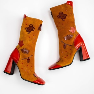Puede incluir: Botas altas de ante amarillo mostaza con detalles rojos. Las botas tienen aplicaciones de hojas y bellotas, puntera cuadrada y tacón ancho. Altura de las botas: aproximadamente 30 cm.