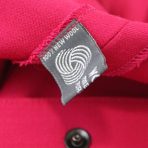 May include: A close-up of a gray fabric label with white text that reads "100% New Wool" and a white wool symbol. The label is attached to a bright pink fabric.