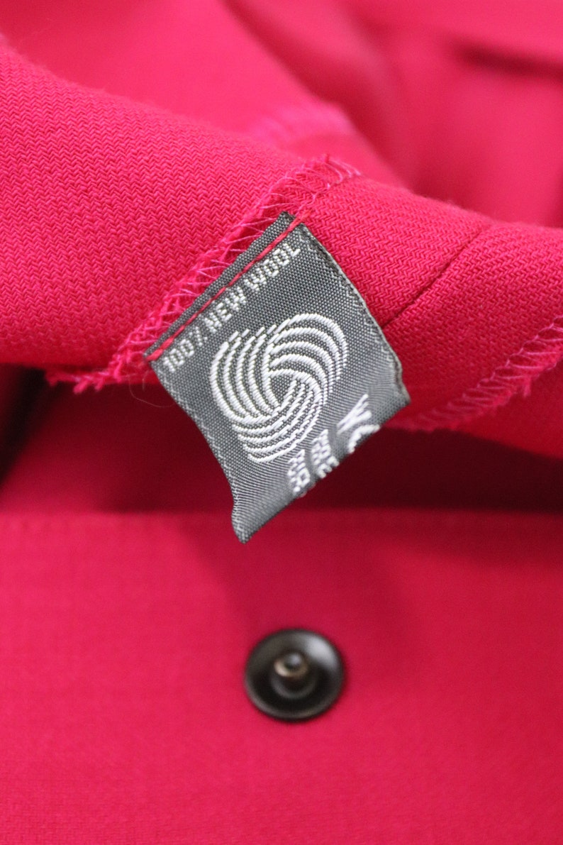 May include: A close-up of a gray fabric label with white text that reads "100% New Wool" and a white wool symbol. The label is attached to a bright pink fabric.