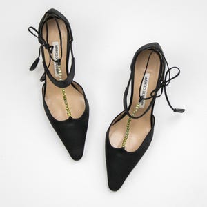 Puede incluir: Un par de zapatos de tac&oacute;n alto de sat&eacute;n negro con punta. Los zapatos presentan un delicado dise&ntilde;o de tiras con una fila de cuentas verdes y el nombre de la marca "MANOLO BLAHNIK" impreso en la tira. Los tacones son finos y los zapatos tienen cierres de lazo.