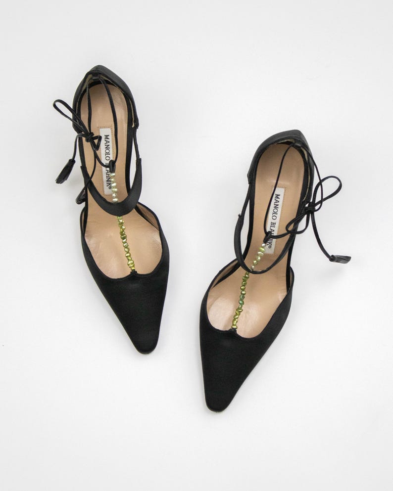 Puede incluir: Un par de zapatos de tac&oacute;n alto de sat&eacute;n negro con punta. Los zapatos presentan un delicado dise&ntilde;o de tiras con una fila de cuentas verdes y el nombre de la marca "MANOLO BLAHNIK" impreso en la tira. Los tacones son finos y los zapatos tienen cierres de lazo.