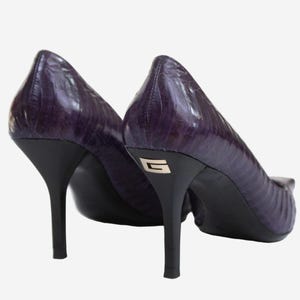 Peut inclure: Une paire d'escarpins à talons hauts violets avec un motif texturé rappelant la peau de reptile. Les chaussures présentent des talons aiguilles noirs, avec un logo en forme de lettre G argenté incrusté à l'arrière de chaque talon, et des semelles noires.