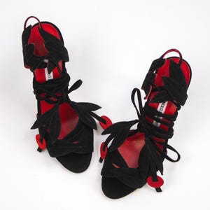 Puede incluir: Sandalias de ante negro con detalles rojos. Las sandalias presentan cortes en forma de hojas y cintas envolventes en el tobillo. Detalles rojos en las suelas y en los extremos de las hojas. Fondo blanco.