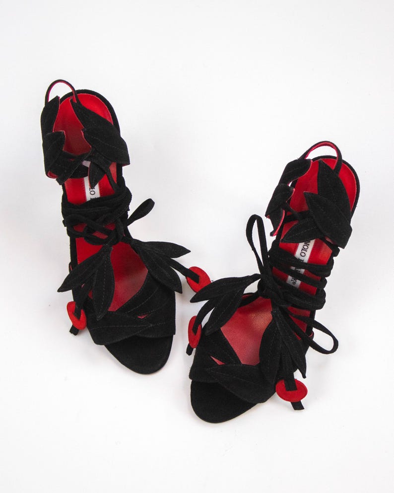 Puede incluir: Sandalias de ante negro con detalles rojos. Las sandalias presentan cortes en forma de hojas y cintas envolventes en el tobillo. Detalles rojos en las suelas y en los extremos de las hojas. Fondo blanco.