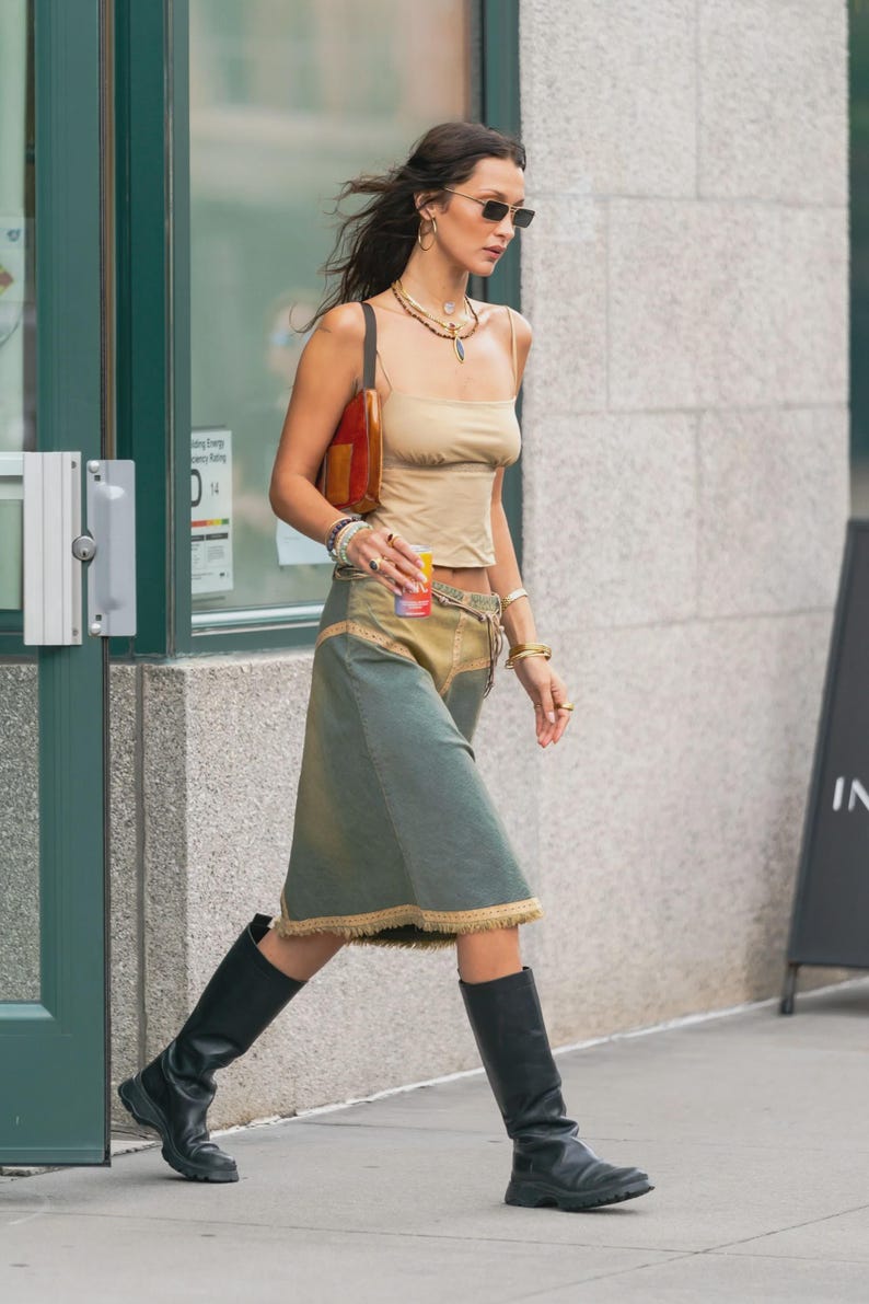 May include: A person wearing a beige tank top, a denim skirt with a frayed hem, and black knee-high boots. Accessories include a brown purse, sunglasses, and layered necklaces. The person is walking on a city sidewalk.