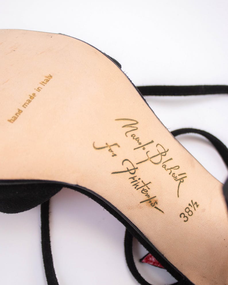 Puede incluir: Sandalia negra de ante con tac&oacute;n alto y fina correa en el tobillo. La suela es beige con letras doradas que dicen "Manolo Blahnik for Printemps" y "hand made in italy". La talla del zapato es 38 1/2.
