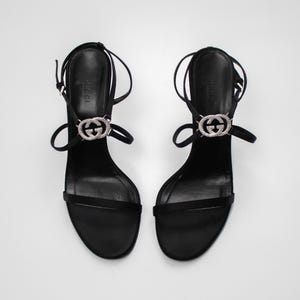 Op de afbeelding: Een paar zwarte sandalen met hoge hakken. Elke sandaal heeft een dun enkelbandje en een bandje over de tenen. De sandalen hebben een zilverkleurig embleem met het logo van het merk. De hakken zijn dun en elegant.