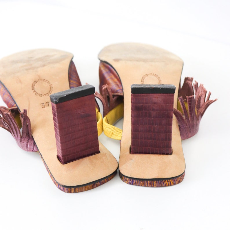 K&ouml;nnte beinhalten: Ein Paar lila und gelbe Sandalen mit Fransen. Die Abs&auml;tze sind klobig und aus dunkelbordeauxfarbenem Material gefertigt. Die Sandalen sind Gr&ouml;&szlig;e 37.
