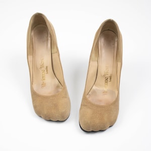 May include: A pair of tan suede high-heeled shoes. The shoes have a rounded toe and a thick heel. The interior of the shoe is a lighter tan color, with the words "Vivienne Westwood London" printed in gold.