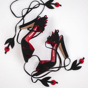 Puede incluir: Un par de sandalias de tac&oacute;n alto de ante negro con plantillas rojas. Las sandalias presentan detalles en forma de hojas y largas correas negras. Detalles circulares rojos se colocan a lo largo de las correas.