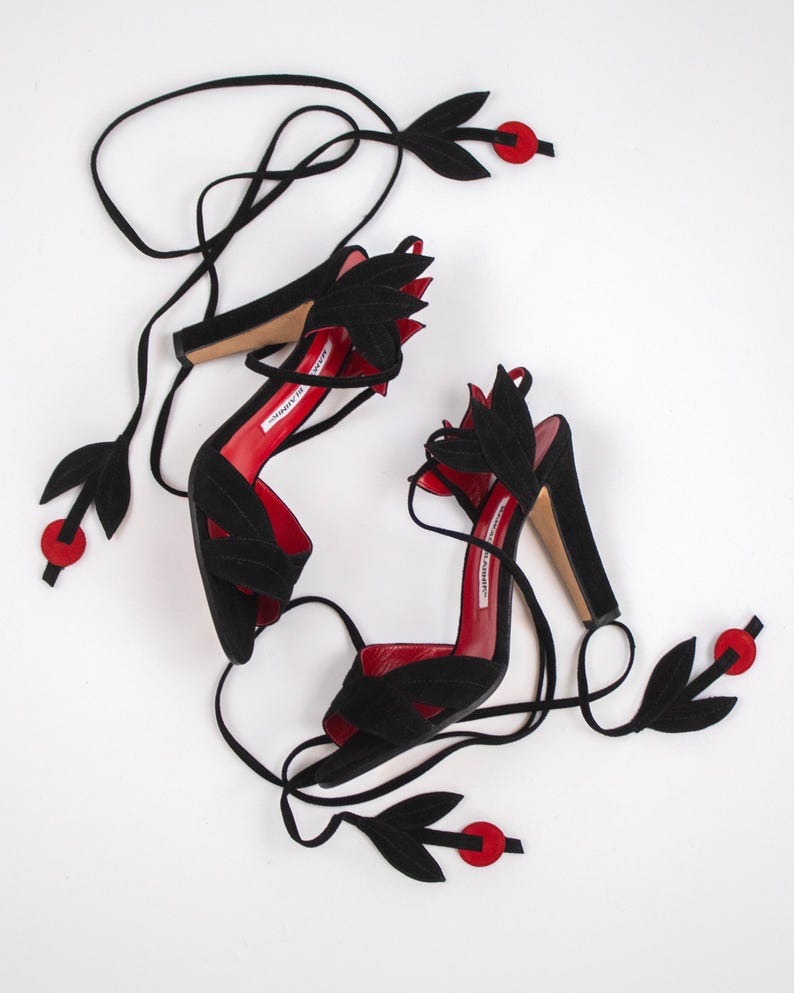 Puede incluir: Un par de sandalias de tac&oacute;n alto de ante negro con plantillas rojas. Las sandalias presentan detalles en forma de hojas y largas correas negras. Detalles circulares rojos se colocan a lo largo de las correas.