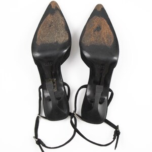 May include: Pair of black high-heeled shoes with pointed toes and ankle straps. The heels are thin and the soles show wear. The shoes are set against a white background.