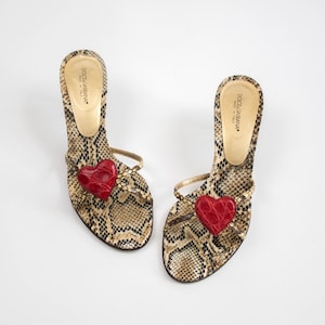 Pode incluir: Sandálias de salto Dolce & Gabbana com padrão de pele de cobra em tons de castanho e bege. Cada sandália tem um detalhe de coração vermelho na tira do dedo. Os saltos são finos e as tiras douradas. Feito na Itália.