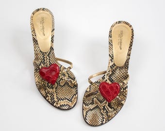May include: A pair of Dolce & Gabbana heeled sandals. The sandals feature a snakeskin pattern in shades of brown and tan, with a red heart embellishment on the toe strap. The heels are thin and the straps are gold.