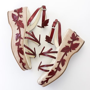 May include: Pair of beige wedge sandals with burgundy leather leaf and vine detailing. The sandals have burgundy straps with silver buckles. The wedge heels are approximately 3 inches high.