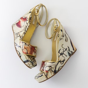 May include: Pair of wedge sandals with floral and abstract print. The cream-colored shoes feature a gold ankle strap and trim. The wedge heel has a height of approximately 4 inches.