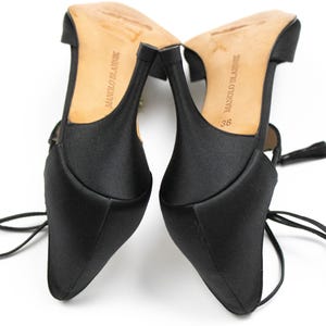 Puede incluir: Par de zapatos de tac&oacute;n alto de sat&eacute;n negro con puntera afilada y tacones finos. Las suelas son de color marr&oacute;n claro con la marca "MANOLO BLAHNIK" impresa. Los tacones miden aproximadamente 7,6 cm. Los zapatos tienen delicados lazos en el tobillo.