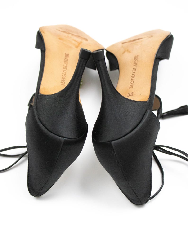 Puede incluir: Par de zapatos de tac&oacute;n alto de sat&eacute;n negro con puntera afilada y tacones finos. Las suelas son de color marr&oacute;n claro con la marca "MANOLO BLAHNIK" impresa. Los tacones miden aproximadamente 7,6 cm. Los zapatos tienen delicados lazos en el tobillo.