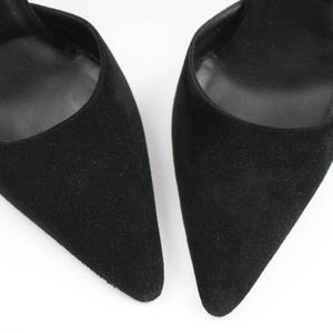 May include: Close-up of two black suede high heels with pointed toes. The shoes have a classic design and a sleek, elegant appearance. The heels are set against a white background.