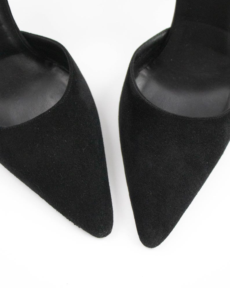 May include: Close-up of two black suede high heels with pointed toes. The shoes have a classic design and a sleek, elegant appearance. The heels are set against a white background.