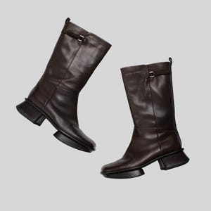 May include: Two brown leather boots with a mid-calf height and a square toe. Each boot has a small buckle detail at the top and a block heel. The boots are set against a plain background.