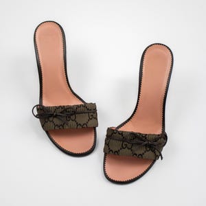 Puede incluir: Un par de sandalias de tacón alto. Las sandalias presentan una plantilla marrón claro, una suela negra y una tira de tela verde oscuro con un patrón repetido. Un pequeño lazo adorna la tira. Los tacones miden aproximadamente 10 cm de alto.