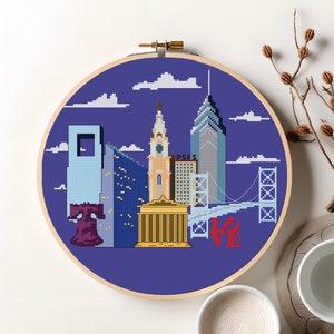 May include: A purple and blue embroidery hoop with a cross-stitch design of the Philadelphia skyline. The skyline includes the Liberty Bell, City Hall, and the Ben Franklin Bridge. The word "LOVE" is stitched in red below the skyline.